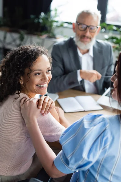 Joyful interracial same sex couple looking at each other near blurred sales manager in real estate agency — Stock Photo