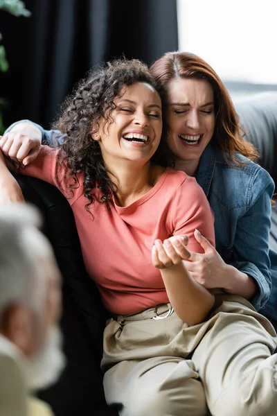 Excitada pareja de lesbianas multiétnicas con los ojos cerrados riendo cerca de psicólogo borroso en la sala de consulta - foto de stock