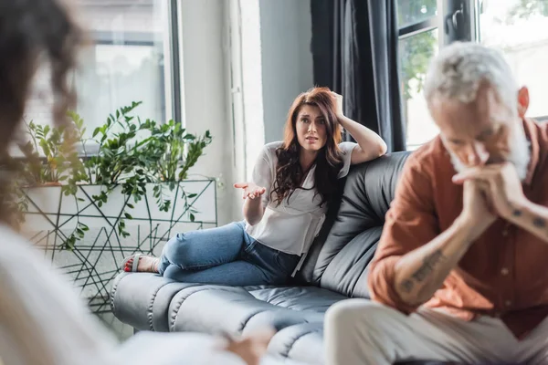 Mujer preocupada hablando con psicólogo borroso mientras está sentado en el sofá cerca de marido deprimido - foto de stock
