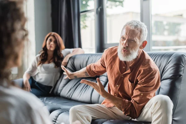 Displeased man pointing at upset wife while talking to blurred african american psychologist — Stock Photo