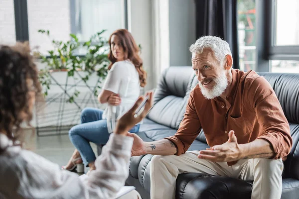 Mann mittleren Alters gestikuliert im Gespräch mit afrikanisch-amerikanischem Psychologen nahe beleidigter Ehefrau — Stockfoto