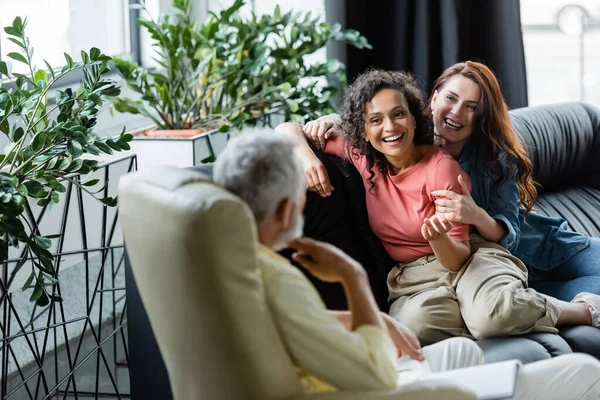 Alegre multiétnico lesbianas sentado en sofá cerca borrosa psicólogo - foto de stock