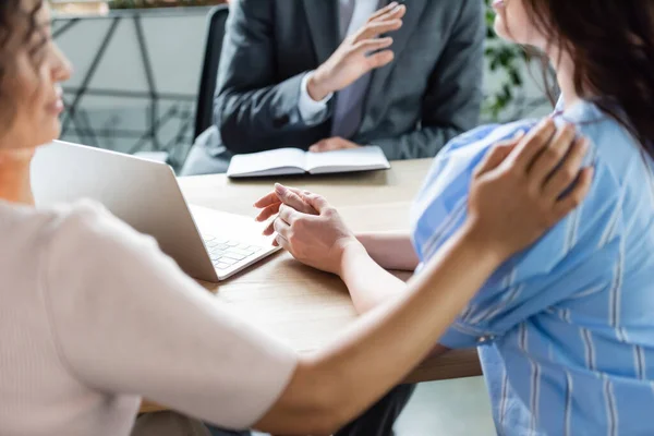 African american woman touching shoulder of lesbian girlfriend near laptop and blurred  broker in real estate agency — Stock Photo