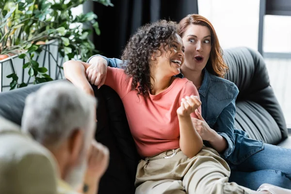 Amazed and cheerful interracial lesbians sitting on couch near blurred psychologist in consulting room — Stock Photo