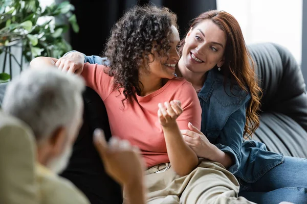 Psicólogo borrosa cerca feliz interracial lesbianas pareja sentado en sofá durante la consulta - foto de stock