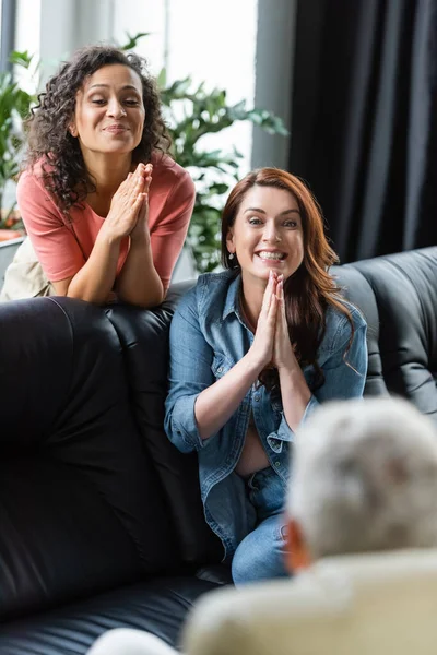 Smiling interracial lesbian couple showing please gesture near blurred psychologist — Stock Photo