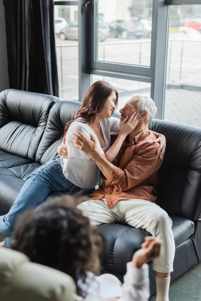 Zufriedenes Paar umarmt sich, während es auf dem Sofa in der Nähe verschwommener afrikanischer amerikanischer Psychologe sitzt — Stockfoto