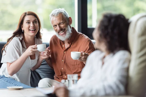 Coppia sorridente seduta con tazze di caffè vicino sfocato psicologo africano americano — Foto stock