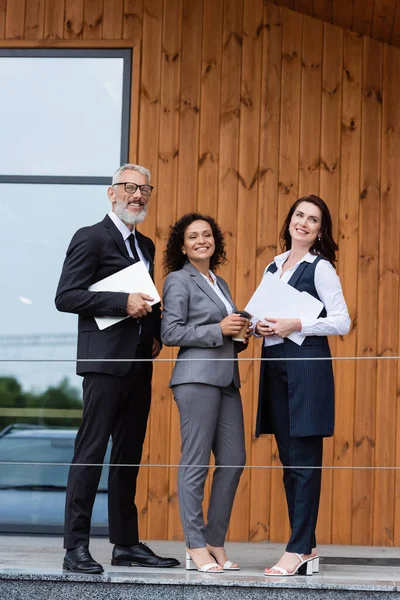 Socios comerciales interracial y corredor de bienes raíces sonriendo cerca de la construcción al aire libre - foto de stock