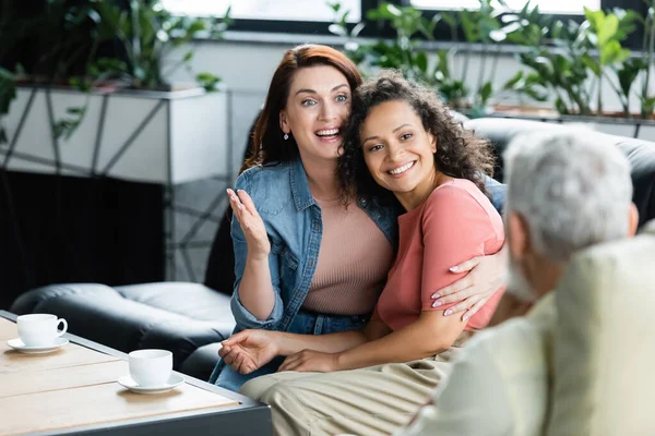 Happy lesbian woman embracing african american girlfriend during consultation with psychologist on blurred foreground — Stock Photo