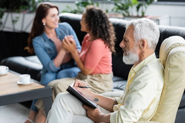 Psicóloga de mediana edad escribiendo en cuaderno cerca de pareja lesbiana interracial cogida de la mano en el sofá - foto de stock