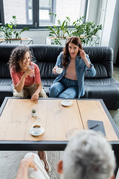 Multiethnic lesbian couple sitting on sofa with water and coffee near blurred psychologist — Stock Photo