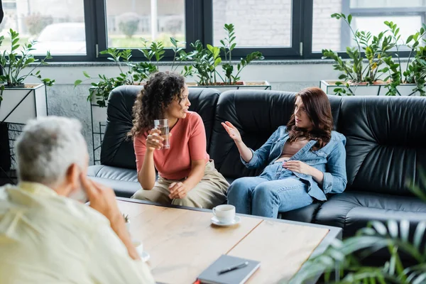 Lesbian woman pointing with hand while talking to african american girlfriend near blurred psychologist — Stock Photo