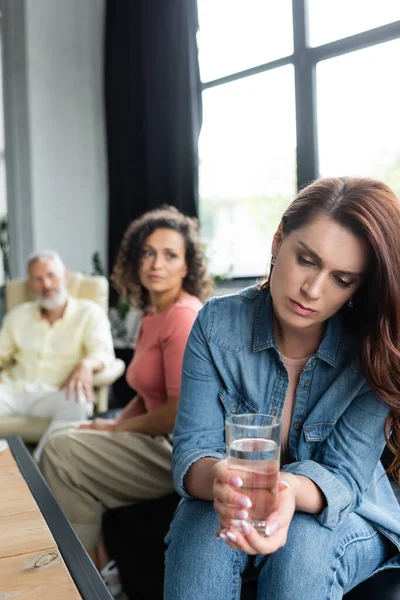 Frustrated woman holding glass of water near african american girlfriend and psychologist on blurred background — Stock Photo