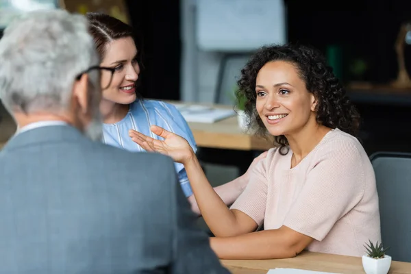 Lächelnde Afroamerikanerin gestikuliert, während sie mit verschwommenem Makler in der Nähe lesbischer Freundin spricht — Stockfoto