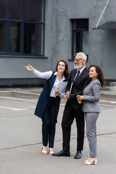 Sorrindo pessoas de negócios multiétnicos olhando para longe perto corretor imobiliário apontando com o dedo ao ar livre — Fotografia de Stock