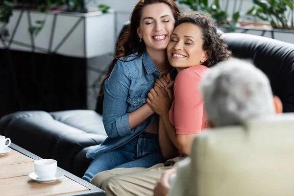 Glückliche multiethnische lesbische Frauen mit geschlossenen Augen, die Händchen halten, während sie auf der Couch neben einem verschwommenen Psychologen sitzen — Stockfoto