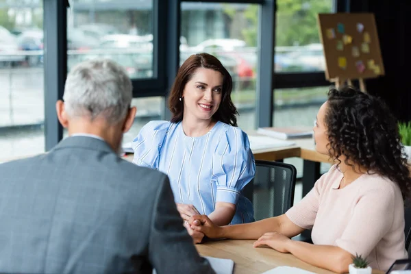 Multiethnisch lesbisches Paar, das einander ansieht und Händchen hält in der Nähe eines verschwommenen Maklers in einer Immobilienagentur — Stockfoto