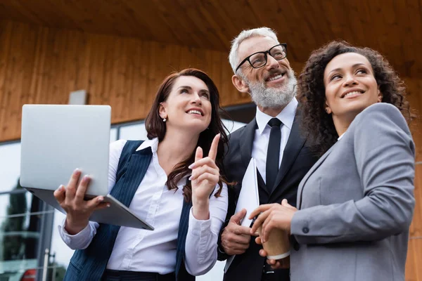 Agente inmobiliario con tableta digital apuntando con el dedo cerca de socios comerciales multiétnicos mirando hacia fuera al aire libre - foto de stock