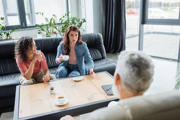 Lesbian woman with cup of coffee talking to blurred psychologist near african american girlfriend — Stock Photo