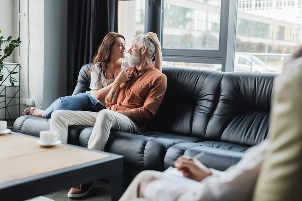 Coppia sorridente che si tiene per mano e si guarda mentre si siede sul divano durante la consultazione psicologica — Foto stock