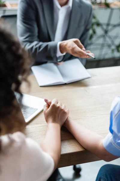 Ausgeschnittene Ansicht eines Maklers, der mit Stift in der Nähe eines leeren Notizbuchs zeigt und ein lesbisches Paar, das die Hände auf verschwommenem Vordergrund hält — Stockfoto