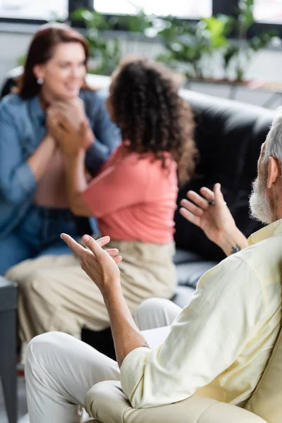 Psicóloga de mediana edad señalando a una pareja de lesbianas multiétnicas cogidas de la mano sobre un fondo borroso - foto de stock