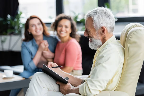 Sorridente psicologo di mezza età che scrive in notebook vicino felice lesbiche interrazziale su sfondo sfocato — Foto stock