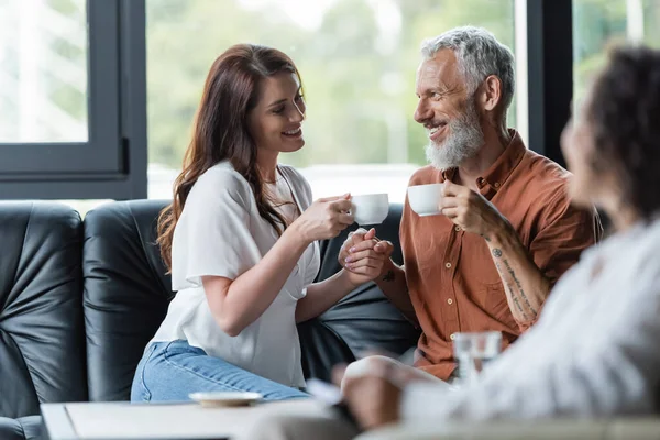 Alegre pareja con tazas de café tomados de la mano cerca borrosa afroamericano psicólogo - foto de stock