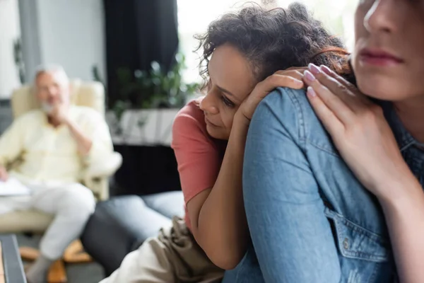 Depressed african american woman leaning on lesbian girlfriend near blurred psychologist — Stock Photo