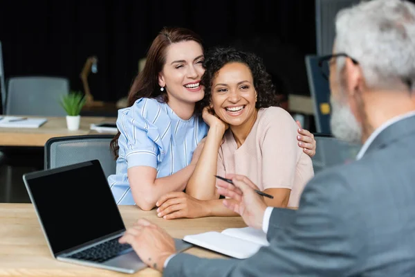 Blurred real estate broker pointing at laptop with blank screen near happy interracial lesbian couple — Stock Photo