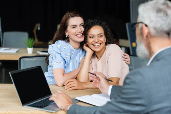 Borrosa agente de bienes raíces apuntando a la computadora portátil con pantalla en blanco cerca de feliz pareja lesbiana interracial - foto de stock