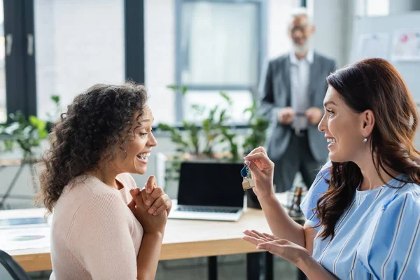 Feliz lesbiana mujer celebración llave cerca sorprendido africano americano novia en real estate agency - foto de stock