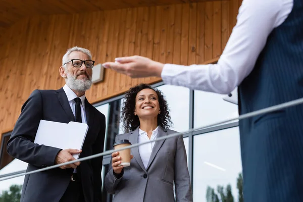 Verschwommener Makler zeigt mit der Hand in die Nähe lächelnder Geschäftspartner mit Ordner und Coffee to go — Stockfoto