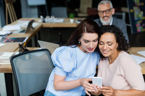 Smiling interracial same sex couple looking at smartphone near real estate sales manager on blurred background — Stock Photo