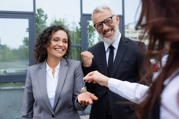 Feliz afroamericana mujer de negocios tomando la clave de borrosa agente de bienes raíces cerca de colega emocionado mostrando gesto de éxito - foto de stock