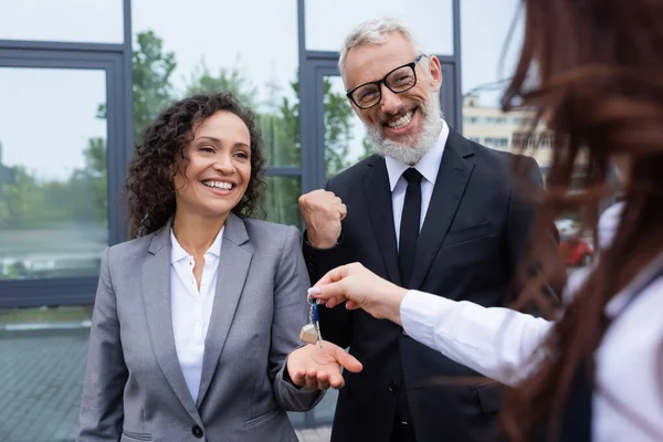 Felice donna d'affari africana americana prendendo la chiave da agente immobiliare offuscata vicino collega eccitato mostrando gesto di successo — Stock Photo