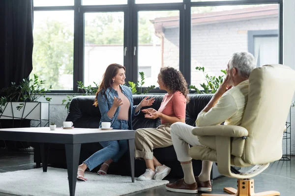 Interracial lesbianas mujeres gesto mientras hablando cerca psicólogo sentado en sillón en consultorio - foto de stock