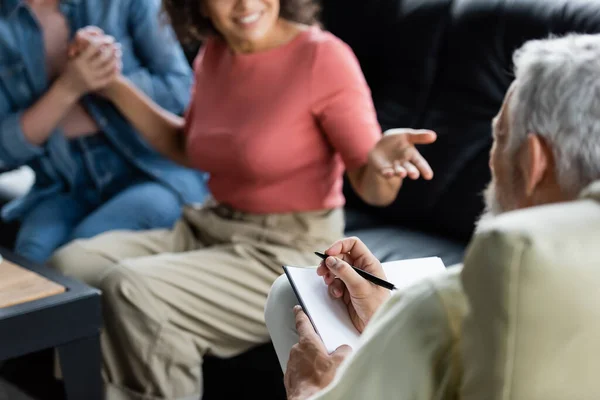 Lächelnde Afroamerikanerin zeigt mit Notizbuch auf verschwommene Psychologin, während sie lesbische Freundin an der Hand hält — Stockfoto