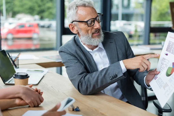 Middle aged realtor pointing at mortgage graphs near interracial lesbian couple holding hands in real estate agency — Stock Photo