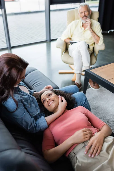 Happy african american woman lying on sofa near lesbian girlfriend and blurred psychologist smiling in armchair — Stock Photo