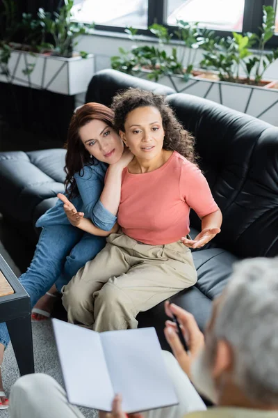 Afrikanerin spricht mit verschwommenem Psychologen mit Notizbuch, während sie auf Sofa neben lesbischer Freundin sitzt — Stockfoto