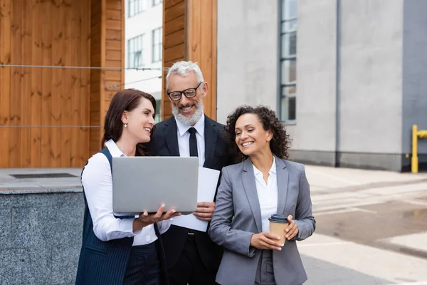 Sorridente agente immobiliare che tiene il computer portatile vicino a felici partner commerciali multietnici all'aperto — Foto stock