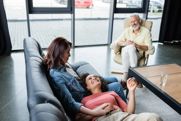 Joyful african american woman lying on couch near lesbian girlfriend holding her hand during visit to psychologist — Stock Photo