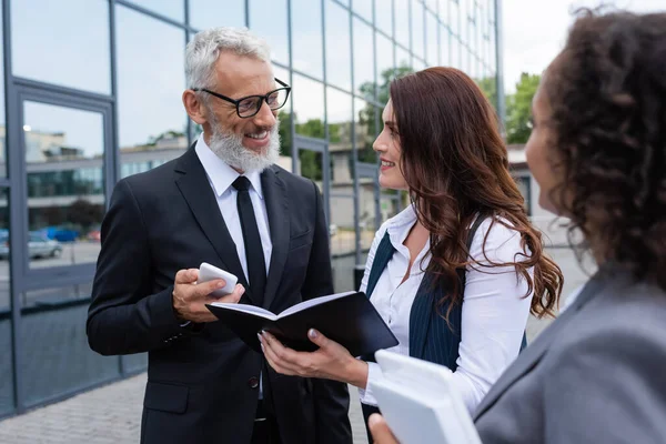 Sonriente hombre de negocios de mediana edad con teléfono inteligente hablando con un agente inmobiliario cerca de un colega afroamericano al aire libre - foto de stock