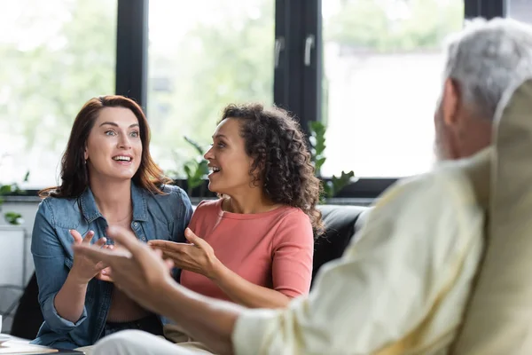 Excited multiethnic lesbian couple gesturing while talking with blurred psychologist during appointment — Stock Photo