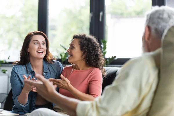 Aufgeregtes multiethnisches lesbisches Paar gestikuliert im Gespräch mit verschwommenem Psychologen während des Termins — Stockfoto