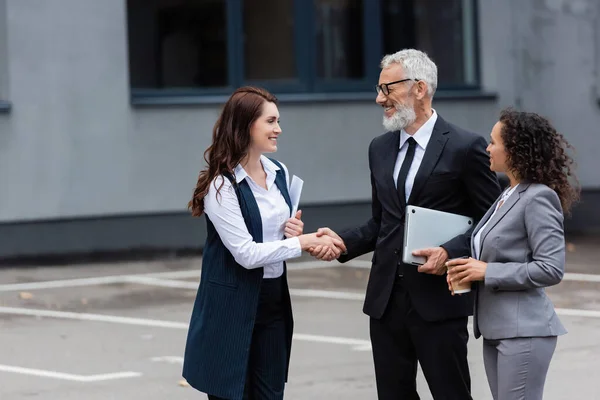 Sonriente hombre de negocios de mediana edad con portátil estrechando la mano con agente inmobiliario cerca de un colega afroamericano - foto de stock