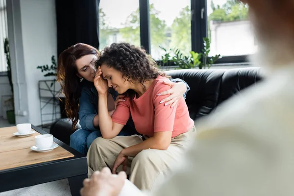 Lesbian woman hugging depressed african american girlfriend near blurred psychologist in consulting room — Stock Photo