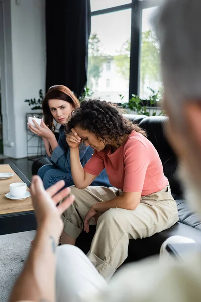 Mujer afroamericana deprimida sentada en un sofá cerca de una novia lesbiana y psicóloga borrosa señalando con la mano - foto de stock